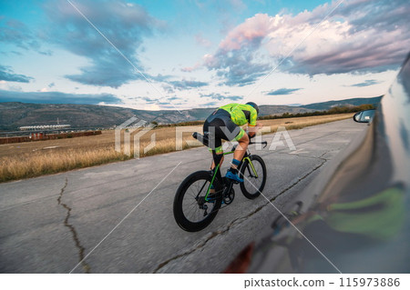 Triathlete riding his bicycle during sunset, preparing for a marathon. The warm colors of the sky provide a beautiful backdrop for his determined and focused effort. 115973886