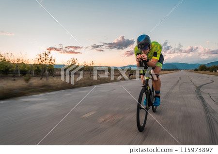 Triathlete riding his bicycle during sunset, preparing for a marathon. The warm colors of the sky provide a beautiful backdrop for his determined and focused effort. Triathlete riding his bicycle during sunset, preparing for a marathon. The warm colors of the sky provide a beautiful backdrop for his determined and focused effort. 115973897