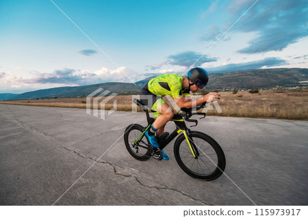 Triathlete riding his bicycle during sunset, preparing for a marathon. The warm colors of the sky provide a beautiful backdrop for his determined and focused effort. Triathlete riding his bicycle during sunset, preparing for a marathon. The warm colors of the sky provide a beautiful backdrop for his determined and focused effort. 115973917