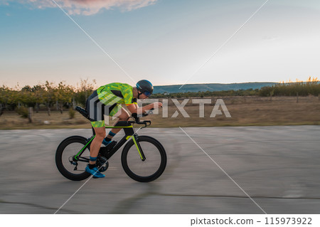 Triathlete riding his bicycle during sunset, preparing for a marathon. The warm colors of the sky provide a beautiful backdrop for his determined and focused effort. 115973922