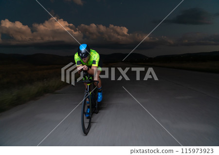 A triathlete rides his bike in the darkness of night, pushing himself to prepare for a marathon. The contrast between the darkness and the light of his bike creates a sense of drama and highlights the A triathlete rides his bike in the darkness of night, pushing himself to prepare for a marathon. The contrast between the darkness and the light of his bike creates a sense of drama and highlights the 115973923