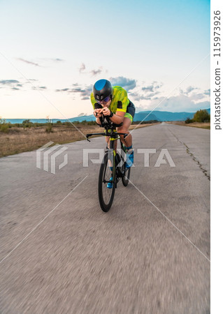 Triathlete riding his bicycle during sunset, preparing for a marathon. The warm colors of the sky provide a beautiful backdrop for his determined and focused effort. Triathlete riding his bicycle during sunset, preparing for a marathon. The warm colors of the sky provide a beautiful backdrop for his determined and focused effort. 115973926