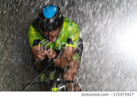 A triathlete braving the rain as he cycles through the night, preparing himself for the upcoming marathon. The blurred raindrops in the foreground and the dark, moody atmosphere in the background add 115973927