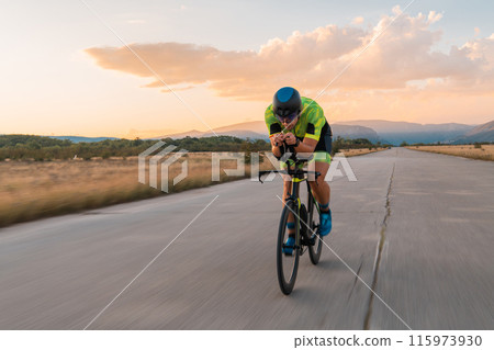 Triathlete riding his bicycle during sunset, preparing for a marathon. The warm colors of the sky provide a beautiful backdrop for his determined and focused effort. 115973930