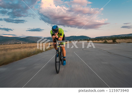 Triathlete riding his bicycle during sunset, preparing for a marathon. The warm colors of the sky provide a beautiful backdrop for his determined and focused effort. 115973940