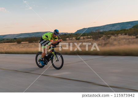 Triathlete riding his bicycle during sunset, preparing for a marathon. The warm colors of the sky provide a beautiful backdrop for his determined and focused effort. Triathlete riding his bicycle during sunset, preparing for a marathon. The warm colors of the sky provide a beautiful backdrop for his determined and focused effort. 115973942