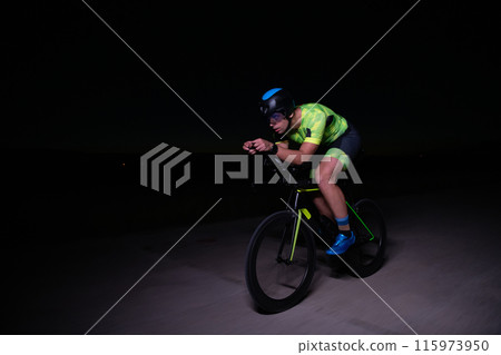 A triathlete rides his bike in the darkness of night, pushing himself to prepare for a marathon. The contrast between the darkness and the light of his bike creates a sense of drama and highlights the 115973950