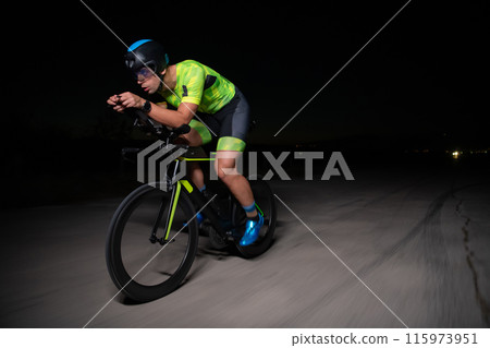 A triathlete rides his bike in the darkness of night, pushing himself to prepare for a marathon. The contrast between the darkness and the light of his bike creates a sense of drama and highlights the 115973951