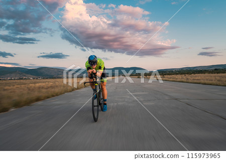 Triathlete riding his bicycle during sunset, preparing for a marathon. The warm colors of the sky provide a beautiful backdrop for his determined and focused effort. 115973965