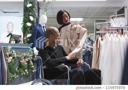 Clothing store assistant talking with arab man customer in wheelchair, examining stylish formal jacket. Shopping mall boutique client with disability receiving advice in choosing woman trendy apparel 115975970
