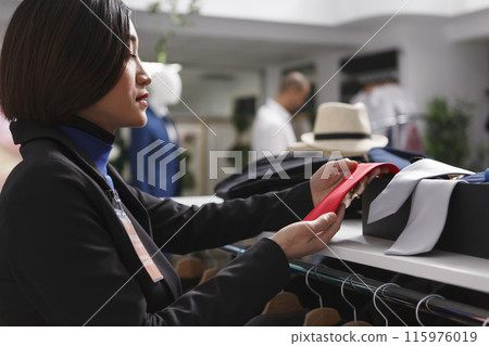 Clothing store asian woman employee showcasing accessory for sale on display shelf. Seller holding red tie while examining merchandise stock in shopping department center boutique 115976019