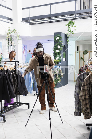 African american social media influencer promoting formal wear collection on smartphone camera in clothing store. Blogger showing jackets while filming live video for followers in boutique 115976043