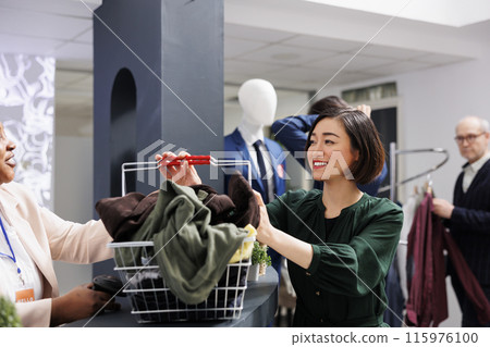 Happy Asian shopaholic woman with shopping basket full of clothes smiling while standing at cash register. Satisfied female customer buying new trendy fashionable clothing items during sales 115976100