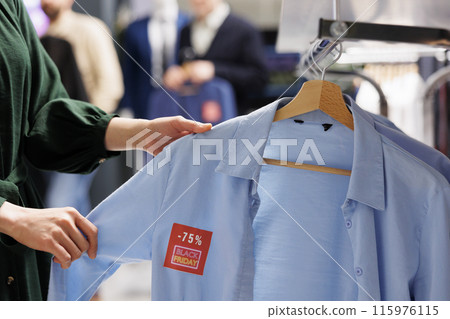 Close up of unrecognizable woman shopper looking at clothes with red discount tag while shopping in outlet store during Black Friday. Female customer holding shirt making purchases in sale season Close up of unrecognizable woman shopper looking at clothes with red discount tag while shopping in outlet store during Black Friday. Female customer holding shirt making purchases in sale season 115976115