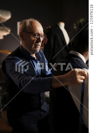 Apprentice suitmaker eager to help master couturier with sartorial measurements for upcoming bespoke fashion collection, close up. Skilled tailor checking handmade suit fitting Apprentice suitmaker eager to help master couturier with sartorial measurements for upcoming bespoke fashion collection, close up. Skilled tailor checking handmade suit fitting 115976116