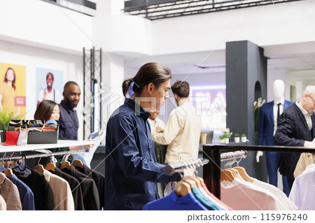Pensive Asian millennial guy customer shopping in apparel store. Confused young man choosing and buying new clothes in shopping mall, looking through male clothing items on hangers Pensive Asian millennial guy customer shopping in apparel store. Confused young man choosing and buying new clothes in shopping mall, looking through male clothing items on hangers 115976240