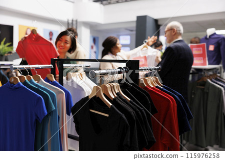 Seasonal sales. Clothes hanging on rack in clothing store with diverse people customers shopping at discounted prices on background. Retail fashion business and bargain hunting concept 115976258