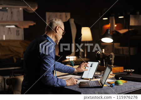 Old experienced suitmaker zooming on tablet, looking at handdrawn sketch designs of sartorial custom made attire. Couturer working on client comission in professional tailoring studio 115976276