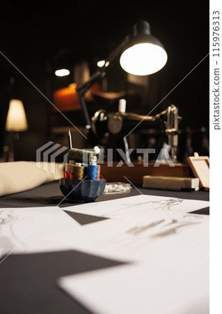 Table full of fashion designs on paper and sewing instruments in tailoring studio. Professional atelier shop workspace with sketch drawings, thread spools, sewing machine 115976313