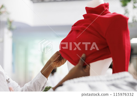 African american woman hands dressing mannequin, showing new arrival in clothing store. Shopping center fashion boutique employee wearing red pullover on dummy model close up African american woman hands dressing mannequin, showing new arrival in clothing store. Shopping center fashion boutique employee wearing red pullover on dummy model close up 115976332