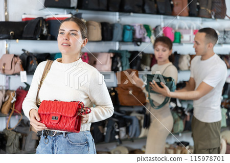 Girl stopped near accessories showcase, holds lady handbag in hands and examines product Girl stopped near accessories showcase, holds lady handbag in hands and examines product 115978701