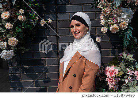 Portrait of a young caucasian woman dressed in a hijab near a wall with flowers. 115979480