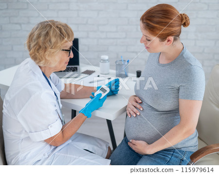 An elderly female doctor checks the blood sugar level of a pregnant red-haired woman. Gestational diabetes. An elderly female doctor checks the blood sugar level of a pregnant red-haired woman. Gestational diabetes. 115979614