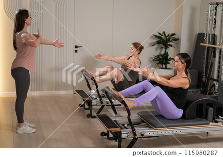 Two pregnant women are doing Pilates on a reformer. The instructor teaches prenatal yoga. Two pregnant women are doing Pilates on a reformer. The instructor teaches prenatal yoga. 115980287