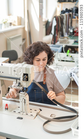 A woman tanner sews a leather belt on a sewing machine. Vertical photo. 115980393