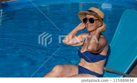 An old woman in a hat and sunglasses is sunbathing on a sun lounger by the pool. Vacation in retirement. An old woman in a hat and sunglasses is sunbathing on a sun lounger by the pool. Vacation in retirement. 115980634