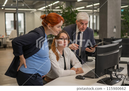 An elderly man, a Caucasian woman and a pregnant woman are discussing work issues at the computer.  115980662