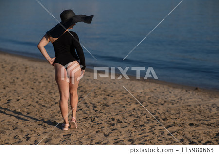 Rear view of woman in large straw hat and black swimsuit posing on the beach. Rear view of woman in large straw hat and black swimsuit posing on the beach. 115980663