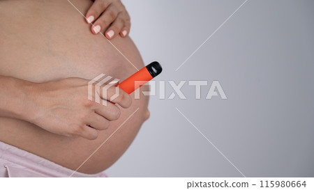 A pregnant woman smokes a vape. A girl holds an electronic cigarette against the background of her bare tummy. Copy space. A pregnant woman smokes a vape. A girl holds an electronic cigarette against the background of her bare tummy. Copy space. 115980664