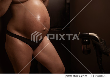 Close-up of the belly of a pregnant woman undergoing an instant tanning procedure. The master applies bronzer with a spray. 115980680