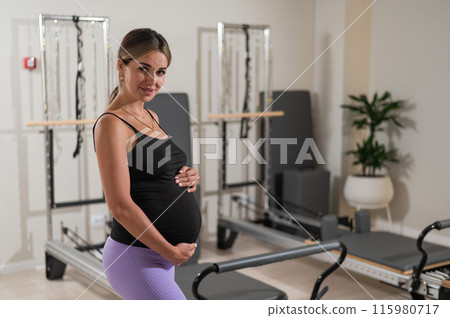 Beautiful pregnant woman stands in a yoga studio and gently touches her belly. Beautiful pregnant woman stands in a yoga studio and gently touches her belly. 115980717