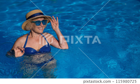 Portrait of an elderly woman in a hat and blue sunglasses swims in the pool.  115980725