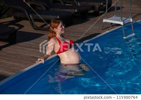 A red-haired pregnant woman in a red bikini is resting in the pool, leaning on the side. 115980764