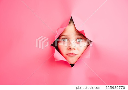 Cute Caucasian girl peeks out of a hole in a paper pink background. 115980770