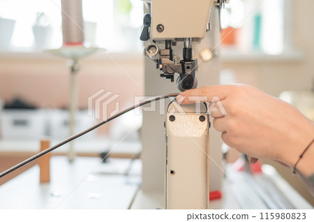 A woman tanner sews a leather belt on a sewing machine. Close-up of hands. 115980823
