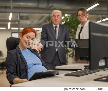 Happy Caucasian pregnant woman sitting at her desk, colleagues gossiping behind her back. Happy Caucasian pregnant woman sitting at her desk, colleagues gossiping behind her back. 115980852
