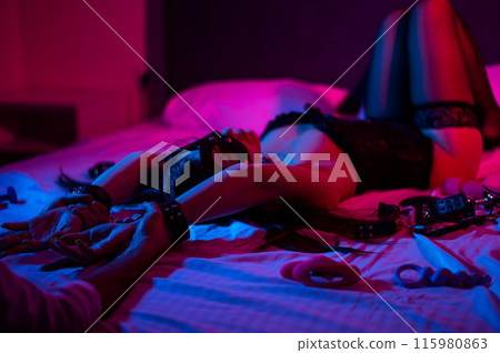 A woman in a leather mask and handcuffs lies on a bed in a blue-red light. Sex toys collection. A woman in a leather mask and handcuffs lies on a bed in a blue-red light. Sex toys collection. 115980863