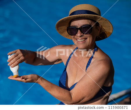 Portrait of an old woman in a straw hat, sunglasses and a swimsuit applying sunscreen to her skin while relaxing by the pool.  115980871