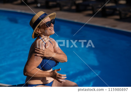Portrait of an old woman in a straw hat, sunglasses and a swimsuit applying sunscreen to her skin while relaxing by the pool.  115980897
