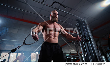 Bald Caucasian bodybuilder training chest using cable crossover in gym. Bald Caucasian bodybuilder training chest using cable crossover in gym. 115980949