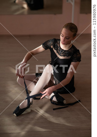 Woman sitting on the floor and tying ribbons on her pointe shoes. Vertical photo.  115980976