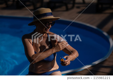 Portrait of an old woman in a straw hat, sunglasses and a swimsuit applying sunscreen to her skin while relaxing by the pool.  115981221