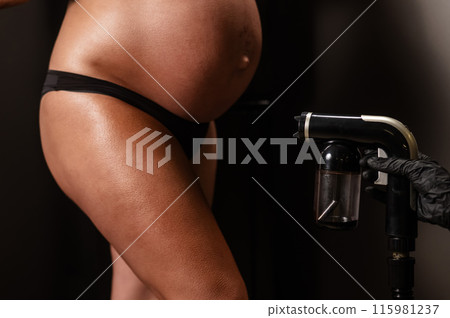 Faceless pregnant woman getting an instant tan. The master applies bronzer with a spray. Faceless pregnant woman getting an instant tan. The master applies bronzer with a spray. 115981237