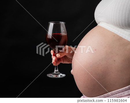 Close-up of the belly of a pregnant woman holding a glass of red wine on a black background. Copy space.  115981604