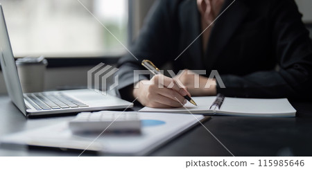 Business Professional Writing Notes at Desk with Laptop and Documents in Modern Office Setting 115985646