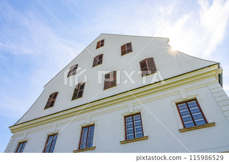 A view of the exterior of the historic manual paper mill building in Velke Losiny, Czechia. The building features a white facade with windows, and the sun is shining through the clouds in the sky. A view of the exterior of the historic manual paper mill building in Velke Losiny, Czechia. The building features a white facade with windows, and the sun is shining through the clouds in the sky. 115986529